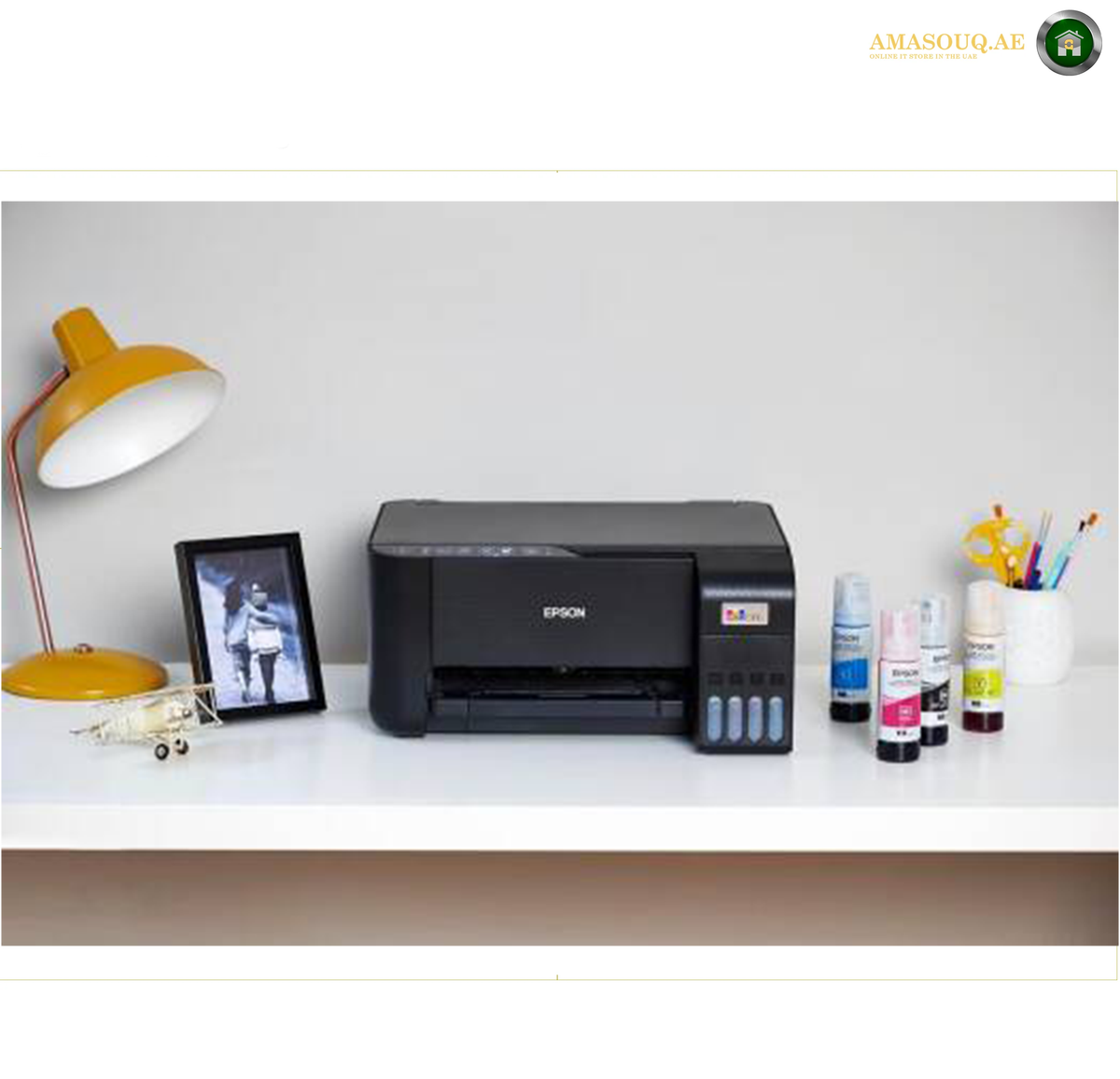 Epson EcoTank L3252 Refillable Four-Colour Ink Tank System | L3252.An Epson inkjet printer sits on a white desk alongside a yellow desk lamp, a framed picture, and several ink bottles against a light gray wall.